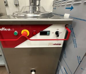 A close-up of a stainless steel commercial kitchen appliance with a red control panel featuring buttons, a digital display, and the brand name “Cattabriga.” Part of another machine is visible to the right.