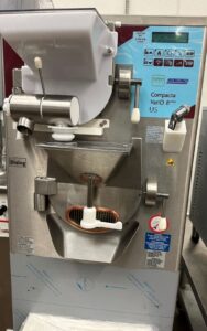 A close-up view of a stainless steel ice cream or gelato machine with labeled buttons, levers, and a chute. The machine has a display panel with blue accents and various operational stickers and symbols.