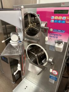 A commercial gelato or ice cream machine with two round metal doors, labeled Compacta Vario 8 by Coldelite, featuring buttons, levers, and a digital display in a stainless steel kitchen setting.