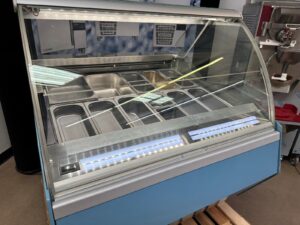 A commercial gelato or ice cream display freezer with an empty glass case and multiple stainless steel containers, sitting on a wooden pallet in a store or shop setting.