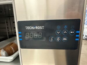 Close-up of a TECH FROST control panel on a stainless steel machine, displaying digital numbers 8.8.8.8 and various buttons for temperature and settings. A wrapped food item is visible on a tray to the left.