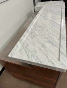 A white marble countertop with gray veining rests on top of a wooden cabinet. The countertop has a raised edge and groove detail, and both pieces are placed against a white wall.