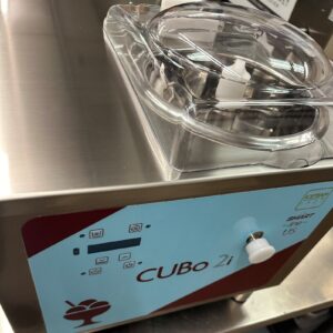 Close-up of a stainless steel ice cream or gelato machine labeled CUBo 2i with a transparent plastic lid on top and control buttons on the front panel.