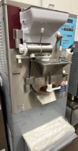 A commercial ice cream or gelato machine with various buttons and levers, a hopper on top, and a dispensing spout in front. The machine is labeled Dialog Compacta Vario 8 US and has a white tray underneath.