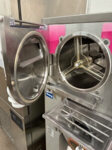 A close-up of a commercial gelato or ice cream machine with its front door open, revealing the mixing chamber and internal metal paddle. The machine is stainless steel with pink accents and a transparent ingredient chute.
