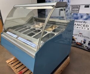 A blue commercial display refrigerator with a curved glass front and multiple stainless steel food pans, placed on a wooden pallet in an indoor setting.