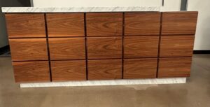 A wooden cabinet with a smooth, glossy finish features a marble top and base. The cabinet has twelve rectangular drawer fronts, displaying a natural wood grain pattern. It stands on a polished concrete floor.