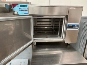 A stainless steel commercial oven with a glass door open, showing empty metal racks inside. The control panel displays buttons and a digital temperature readout. The oven sits on a metal countertop in a clean kitchen.
