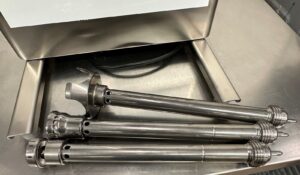 Three stainless steel immersion blender shafts with attachments resting on a metallic countertop beneath a large kitchen appliance.