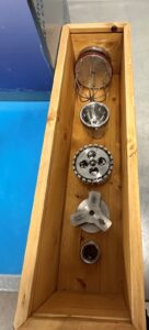 A wooden box holds five metal machine parts arranged in a row, including a wire whisk attachment, a metal bowl, a toothed gear, a three-blade piece, and a small cylindrical component.