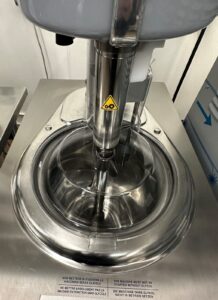 Close-up of a stainless steel industrial machine with a transparent lid, mixing or churning contents inside. A yellow caution sticker is visible on the central spindle and warning instructions are printed on the metal surface nearby.