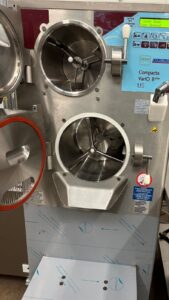 A close-up of an industrial ice cream or gelato machine with two open stainless steel mixing chambers, control panel, and various buttons and levers visible.