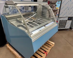 A blue commercial display freezer with a curved glass front, showing several empty metal trays inside, is placed on a wooden pallet in an indoor setting. Another industrial machine is visible in the background.