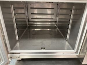 The image shows the inside of a clean, empty stainless steel commercial warming or holding cabinet with metal racks on each side and a drain hole on the bottom.