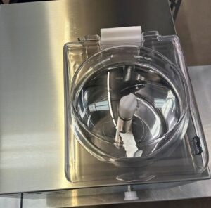 Top-down view of a clean, empty commercial ice cream or gelato maker with a transparent lid, metal paddle, and stainless steel surface reflecting lights above.