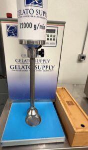 A large gelato machine labeled GELATO SUPPLY and 12000 g/min with a metal mixing tool stands on a blue mat next to a wooden box on a countertop in a clean, industrial kitchen setting.