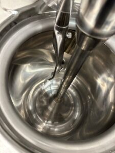 Close-up view of a shiny, stainless steel industrial mixing machine with a central rotating blade inside a cylindrical vessel. The reflective metal surfaces and mechanical parts are clearly visible.