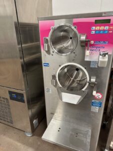 A commercial Coldelite Compacta Vario 8 US machine with two large stainless steel mixing chambers and a control panel, used for making gelato or ice cream, stands in a clean industrial kitchen setting.