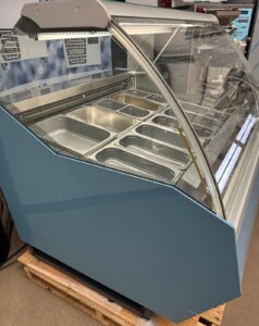 A blue refrigerated display case with a curved glass front, containing several empty metal food trays, is positioned on a wooden pallet in an indoor setting.