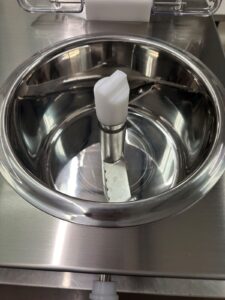Close-up of a shiny stainless steel machine part with a central white plastic component and a metal blade, possibly part of a food processing or mixing device. Reflections are visible on the metallic surface.