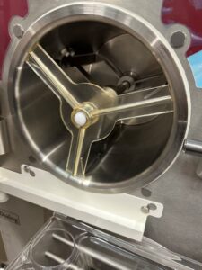 Close-up of the interior of a commercial soft serve or gelato machine, showing a clear plastic mixing paddle inside a round stainless steel chamber.