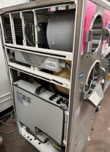 Open side view of a large stainless steel laboratory or medical machine exposing internal components, including wiring, rollers, and a control panel. Pink label and a Dialysis sticker are visible on the exterior.