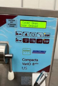 Close-up of an ice cream machine display reading Cover Open Door Opened with various control buttons, warning labels, and part of a dispenser handle visible. The machine is labeled Compacta VariO 8 elite US.