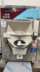 A stainless steel Cattabriga Stargel 4 US gelato machine with a digital display, white plastic hopper, control buttons, and warning labels, situated on a countertop in an industrial kitchen setting.