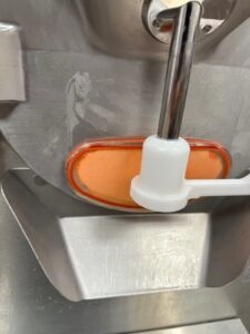 A close-up of a metal machine with a transparent orange cover, a metal rod, and a white plastic handle, likely part of an industrial or kitchen appliance.