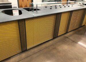 A modern food service counter with a gray countertop and yellow mesh paneling, featuring multiple circular food wells with black lids under a glass sneeze guard, set on a polished concrete floor.