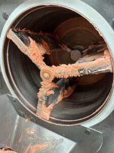 Close-up of a commercial mixer with metal blades covered in thick, pink paste. The cylindrical bowl has remnants of the paste smeared along the sides.