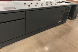 A large, industrial-style black kitchen island with induction burners on top, set on a polished concrete floor in a commercial or institutional setting.