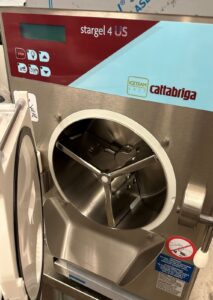 A close-up of a CattaBrega Stargel 4 US ice cream machine with the front door open, revealing the internal mixing chamber and paddle. Control buttons and a digital display are visible on the top panel.