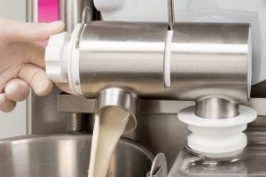 A gloved hand turns a dial on a stainless steel machine as creamy liquid dispenses from a spout into a metal container below.