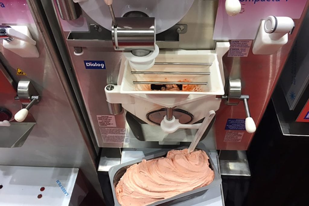 A commercial ice cream machine dispenses pink gelato into a rectangular metal tray below, with a plastic spatula resting in the gelato for serving.