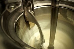 Close-up of an industrial ice cream machine mixing a creamy white mixture with a metal paddle inside a stainless steel container.