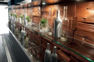 A modern glass shelf against a textured wooden wall holds clear and metallic bottles, glassware, and small potted succulents in glass containers, creating a stylish, decorative display.